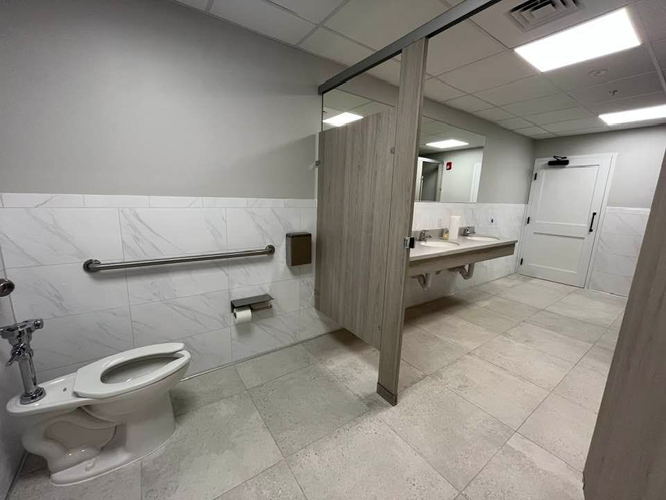 Accessible public restroom with a toilet, sink, and grab bar. Light gray walls, neutral flooring, and wood stall doors.