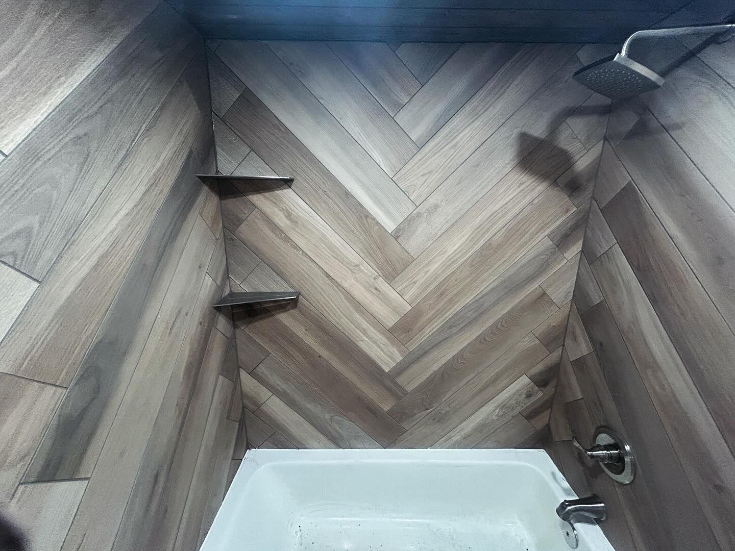 Herringbone wood-look tiled shower with a white bathtub and two shelves. A shower head is visible.