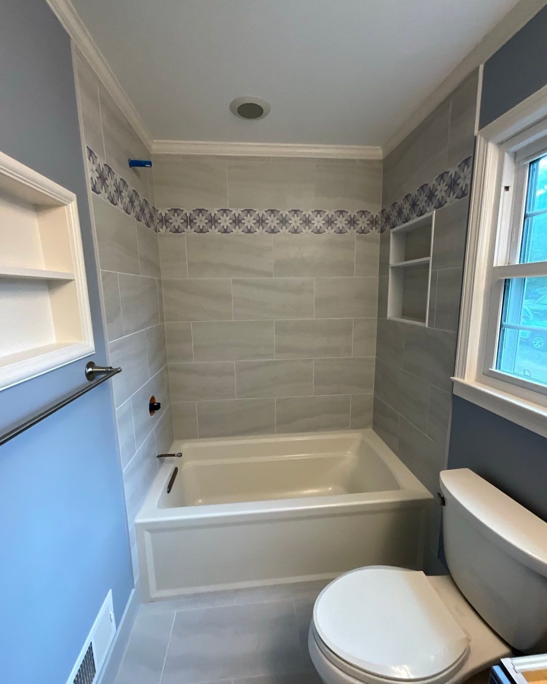Bathroom with light gray tiled shower and tub, blue walls, white trim, toilet, and a window.
