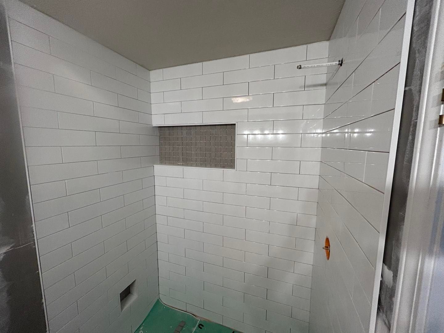 White tiled shower with built-in shelf; partially finished, green floor, door on the right.