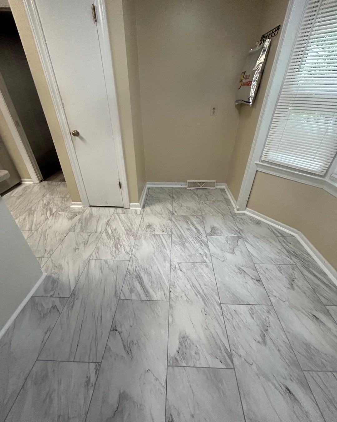 Room with gray and white marble-patterned flooring, white door, beige walls, and a window with blinds.