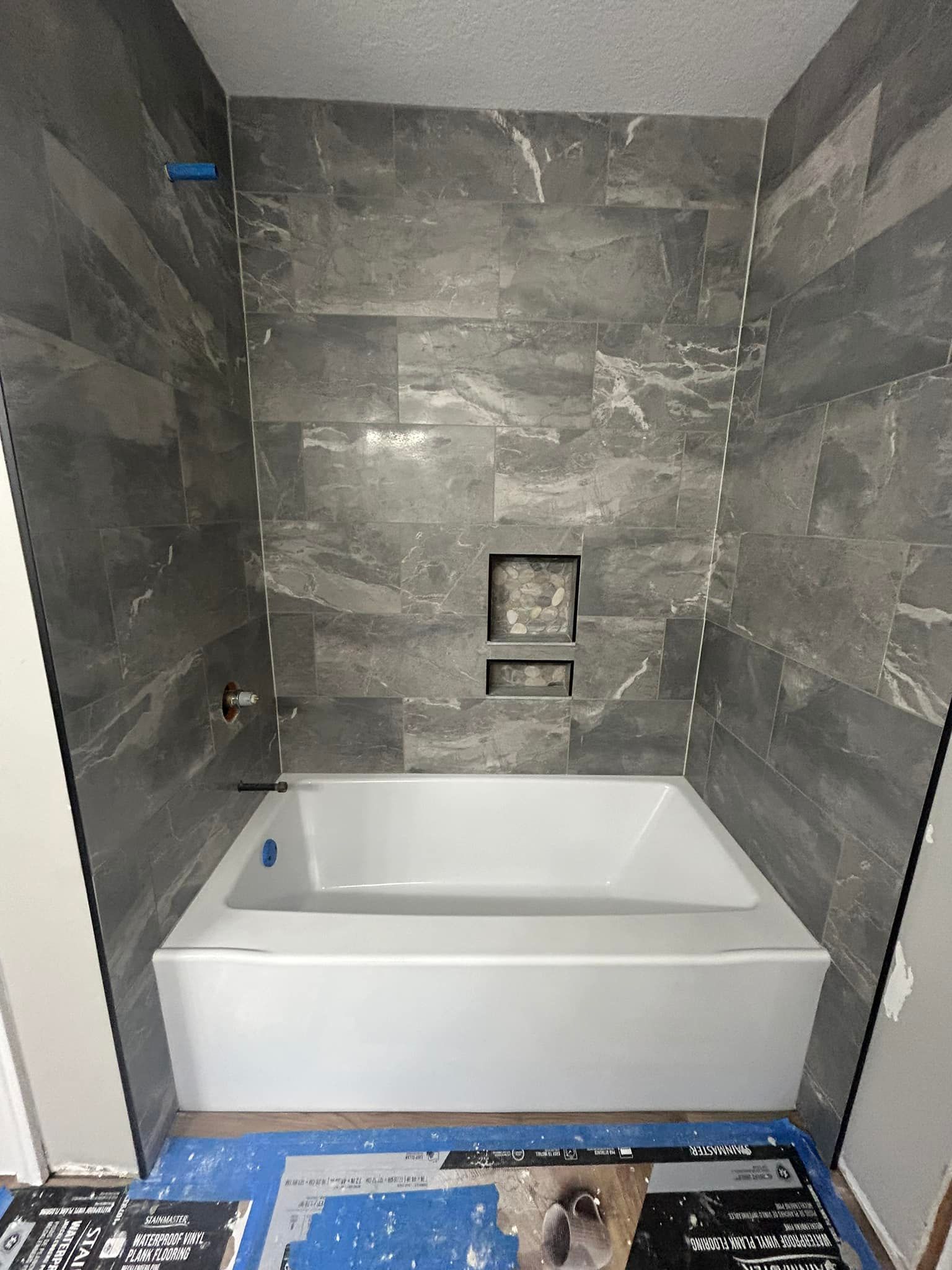 Bathroom with gray tiled walls, white tub, and a built-in shelf.