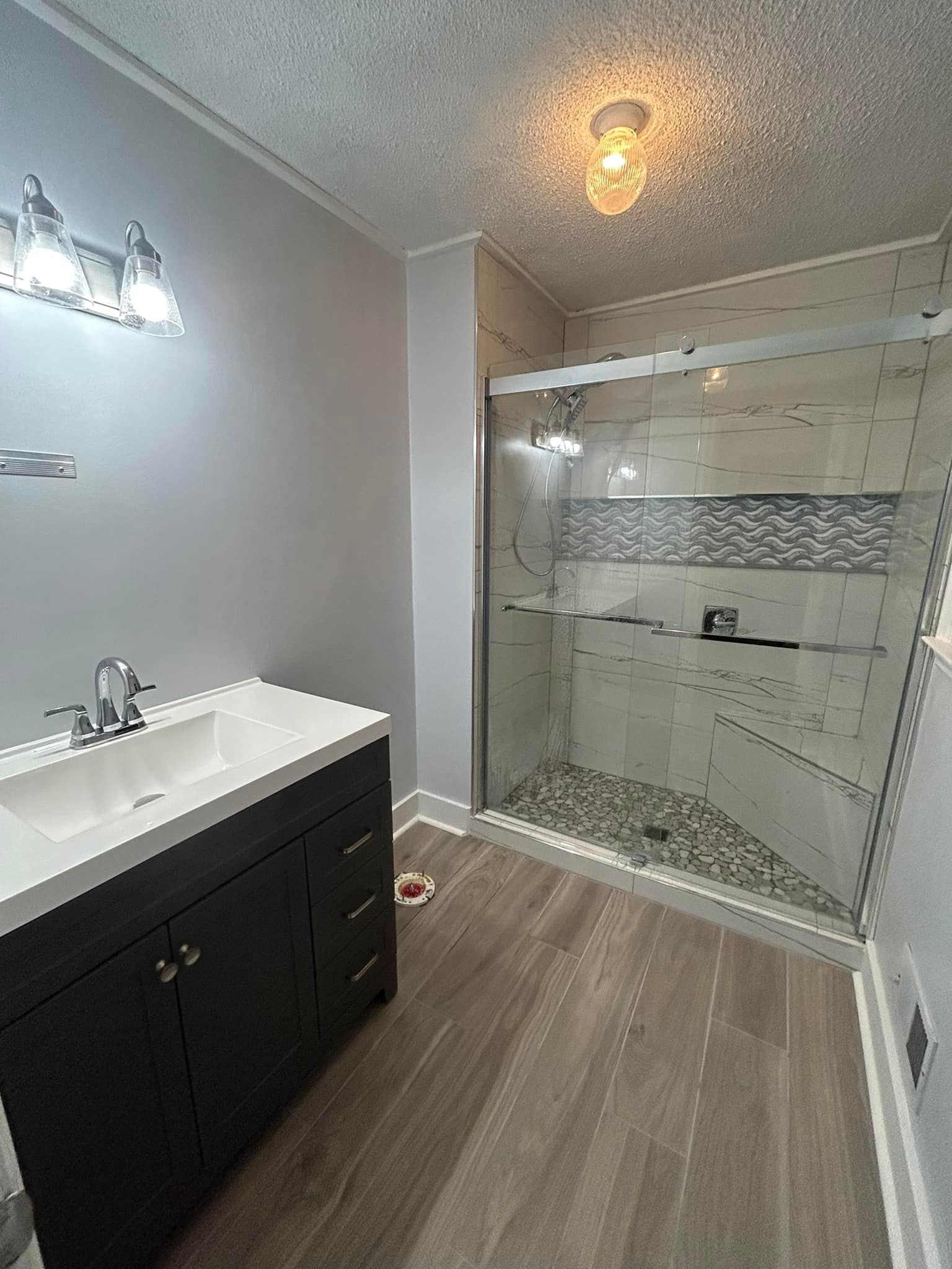 Bathroom with dark vanity, glass shower, light gray walls, and wood-look flooring.