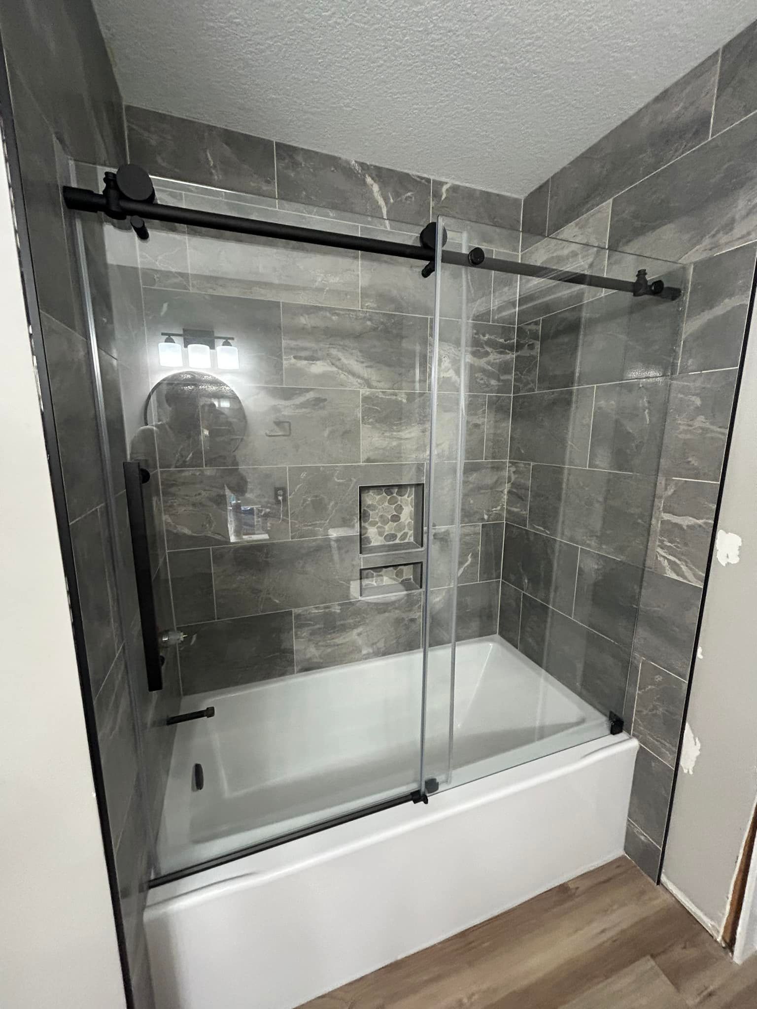 Gray tiled bathroom with a white tub and a black-framed sliding glass shower door.
