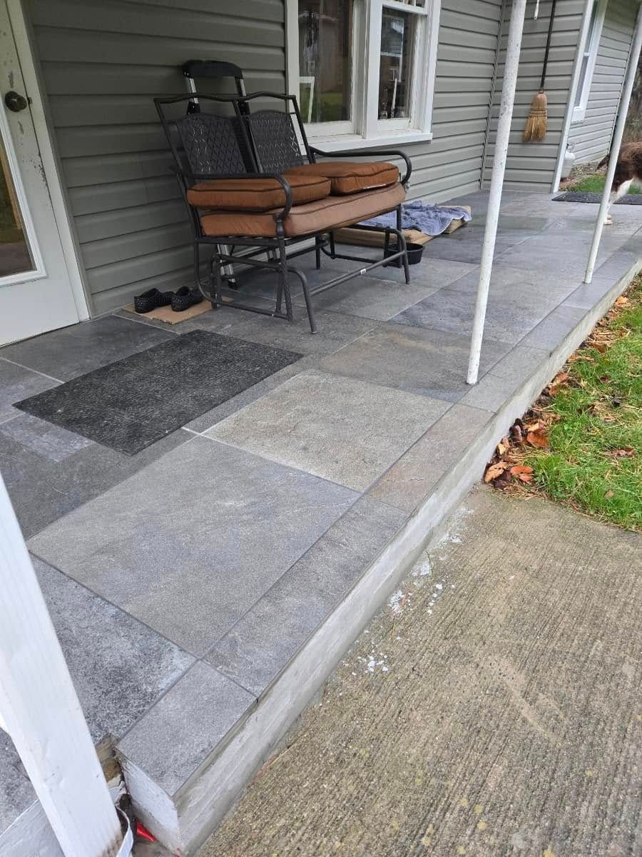 Porch with gray tile flooring, a bench, and a welcome mat. The porch has a concrete border and pillars.