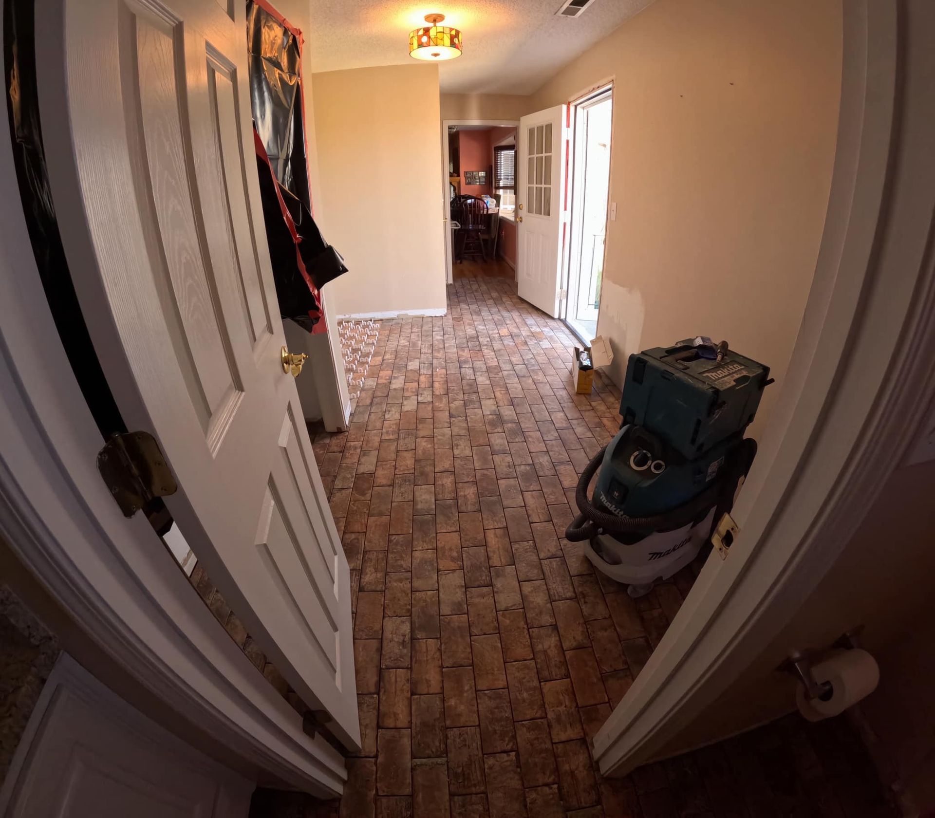 A hallway with brick-like flooring. A door is ajar, showing a view inside the home with a person standing.
