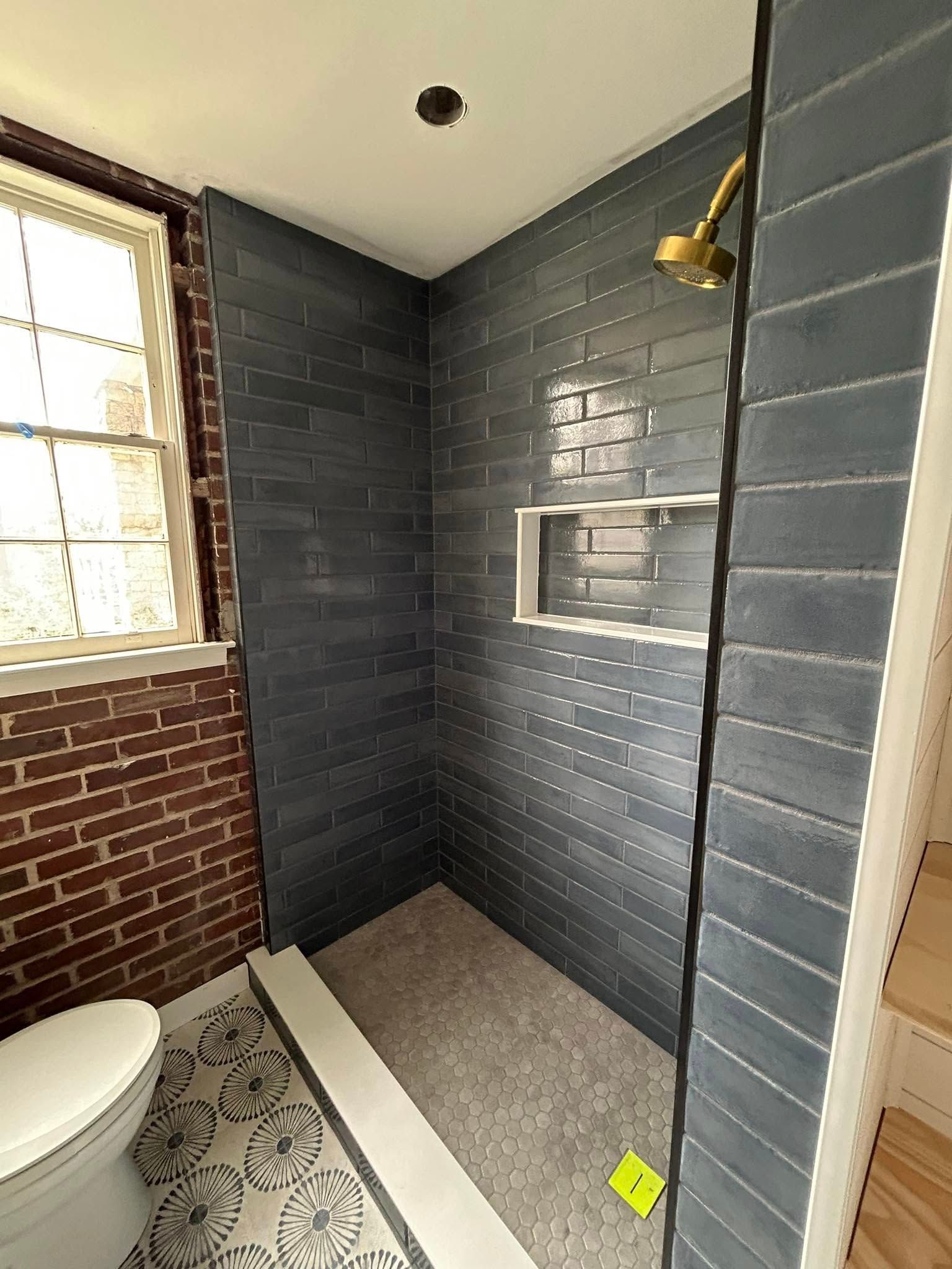 Small shower with blue tile walls, a built-in shelf, and gold showerhead. Toilet and brick wall are also visible.