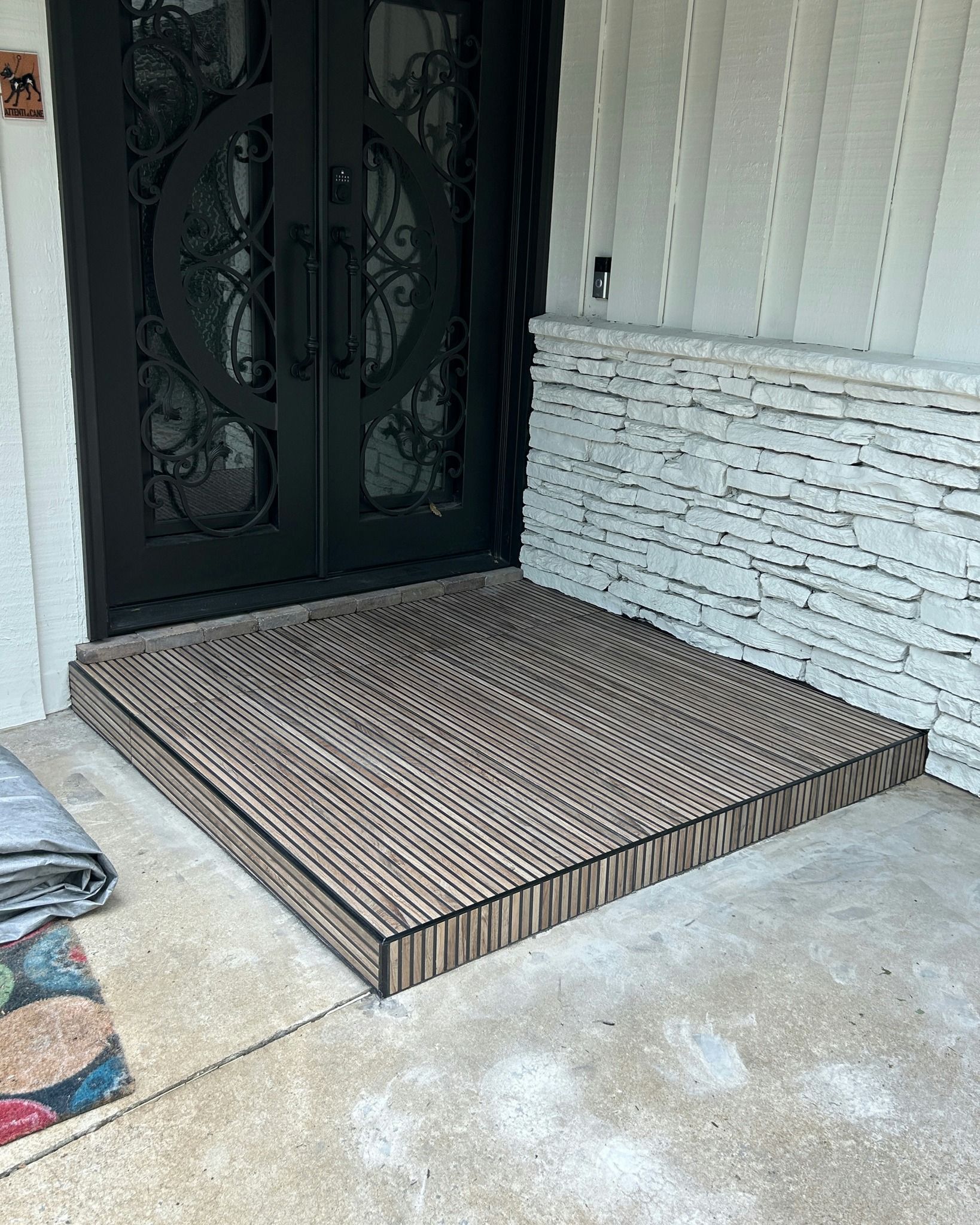 Black door with a brown wooden ramp; gray concrete flooring. A colorful rug is to the left.