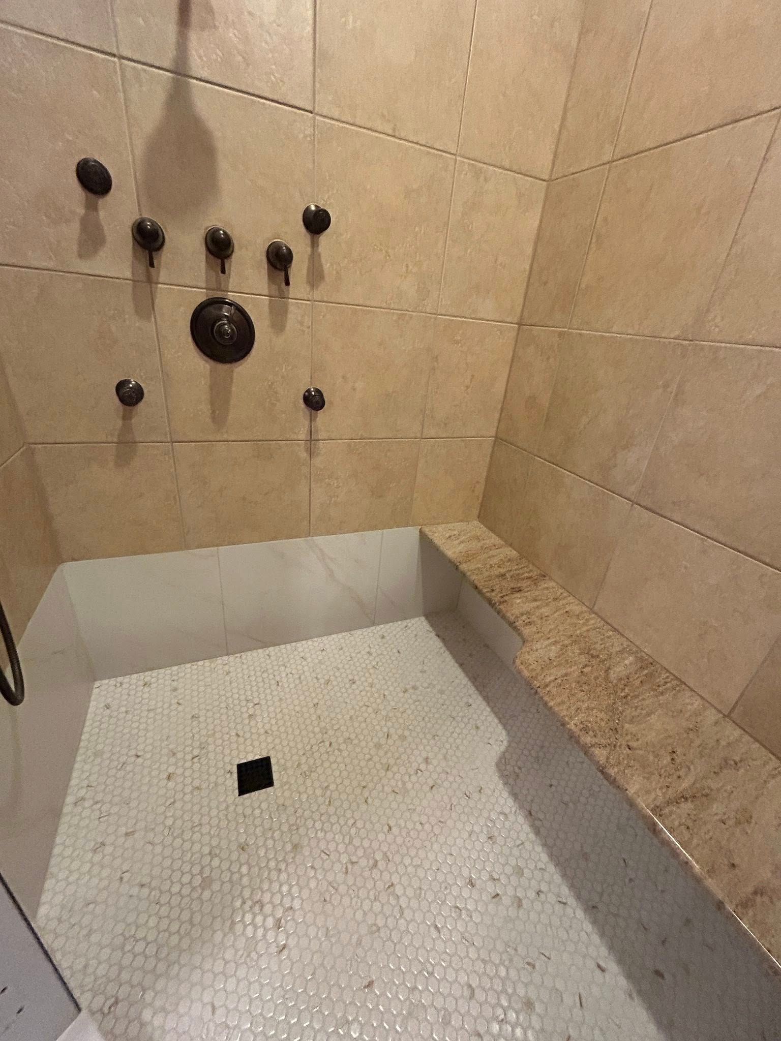 Shower with beige tile walls, pebble floor, and bench. Bronze showerheads and faucet are installed.
