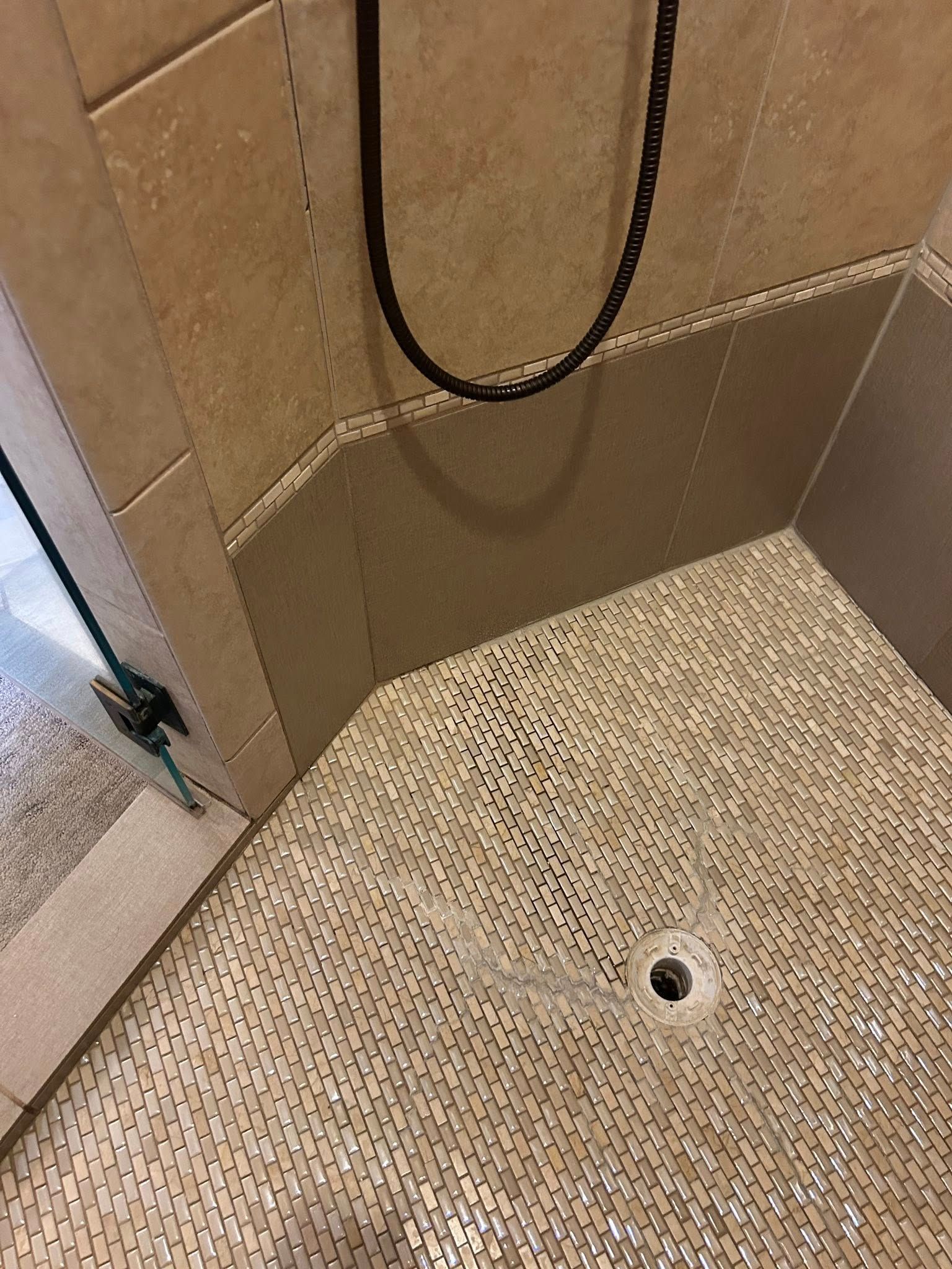 Shower stall with beige tile walls and mosaic floor. Black shower head and drain visible.