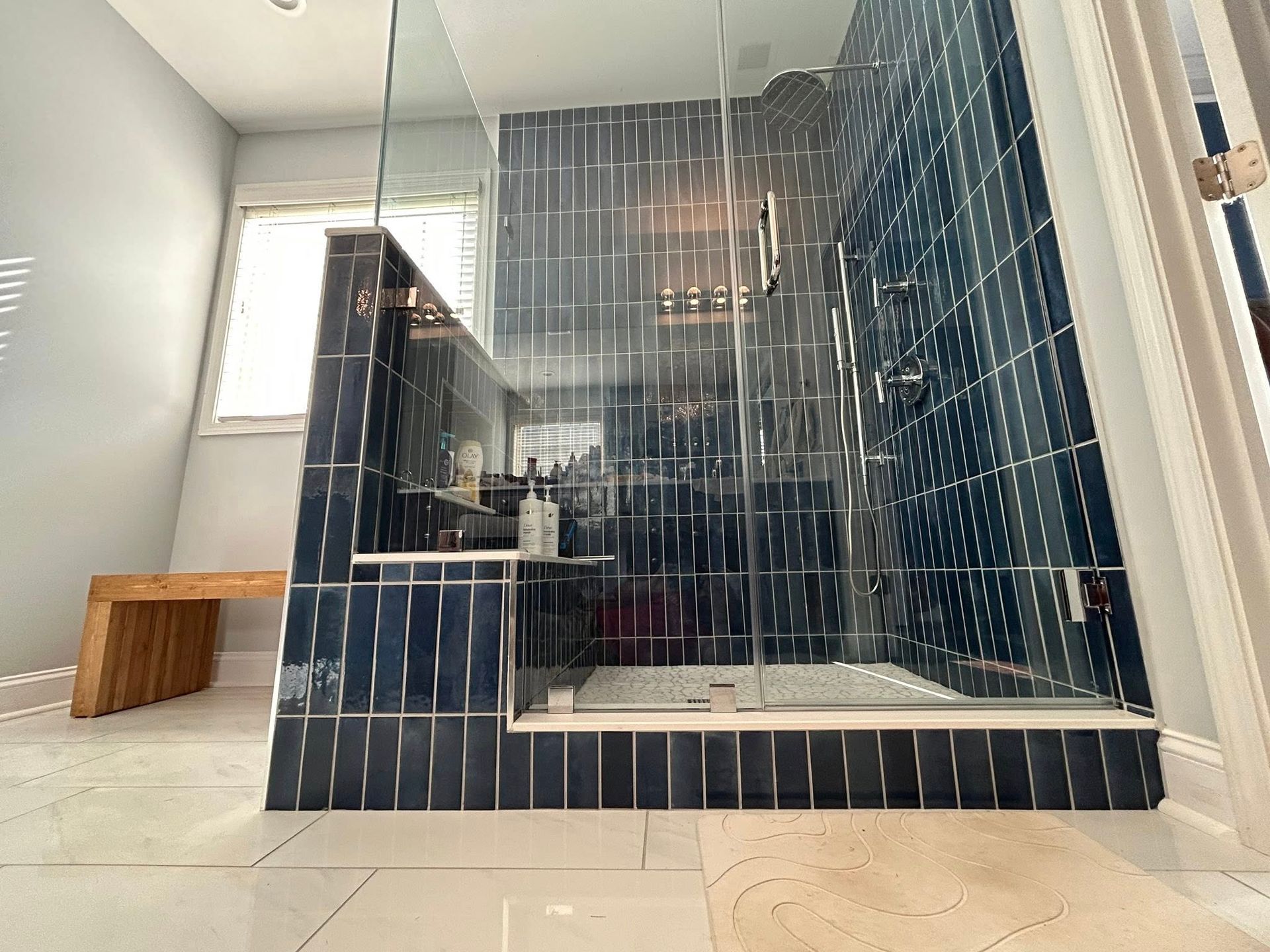Blue-tiled shower with glass doors, white floor, and wooden bench in bathroom.