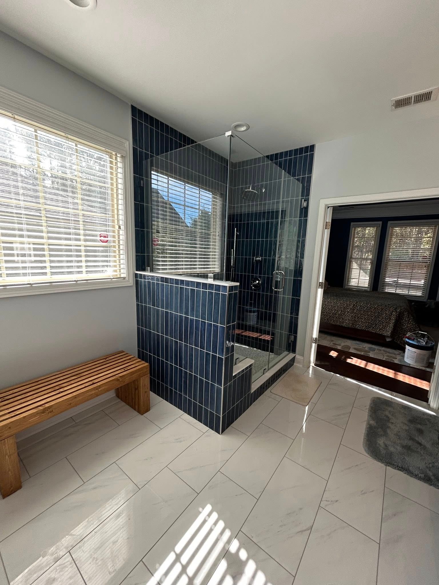 Modern bathroom with blue-tiled shower, white tile floor, wooden bench, window with blinds, and an open doorway.