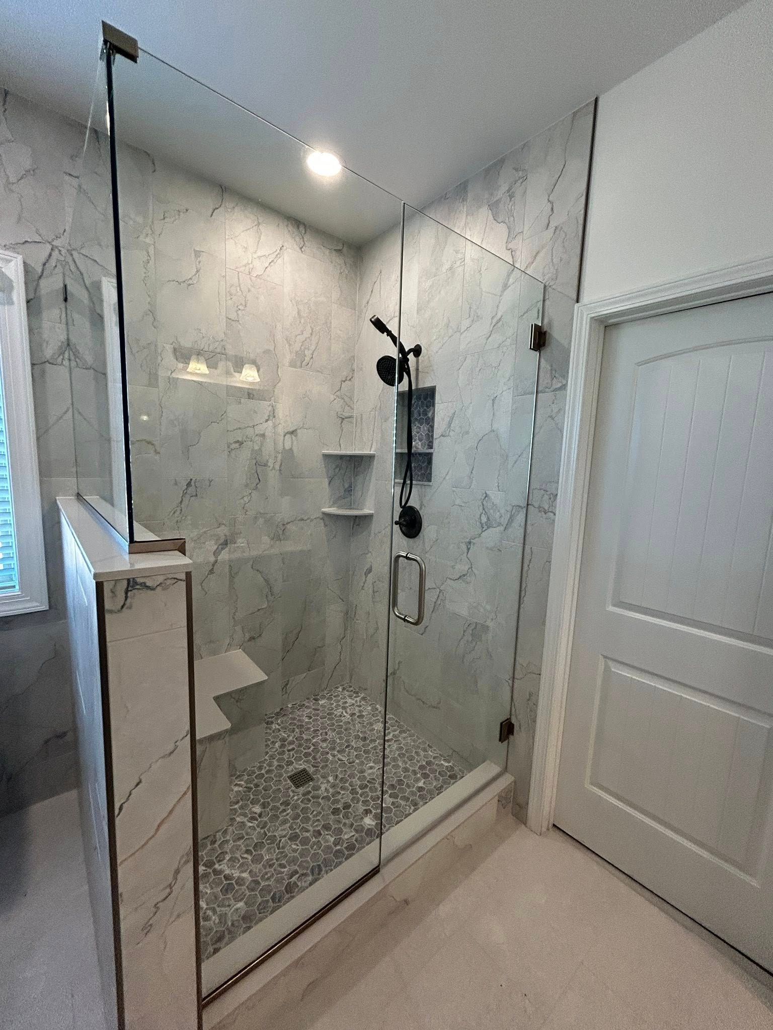 Glass shower with pebble floor, gray marble walls, black fixtures, and white door.