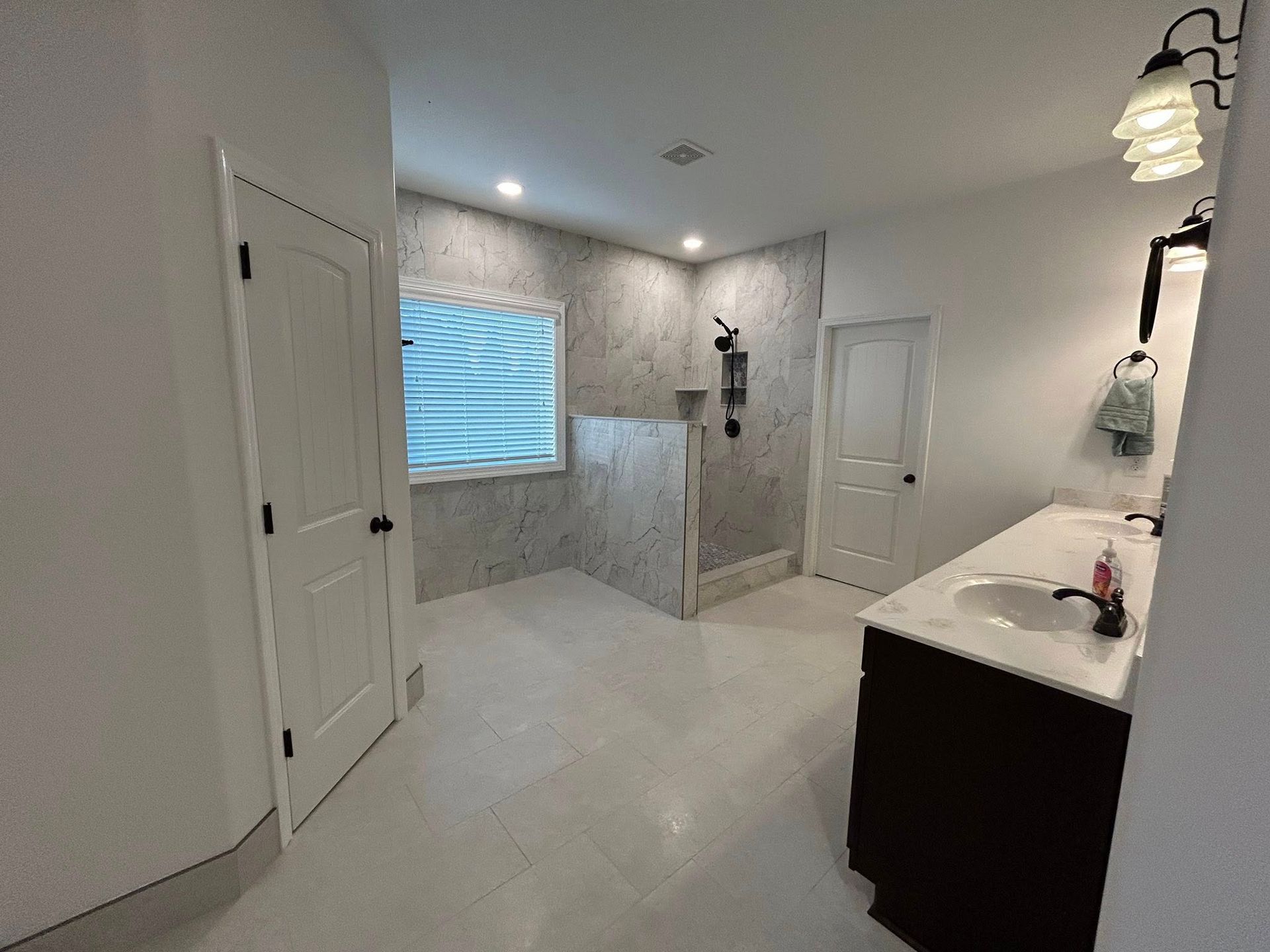 Spacious bathroom with white tile floor, vanity, shower stall, and a closed door on the left.