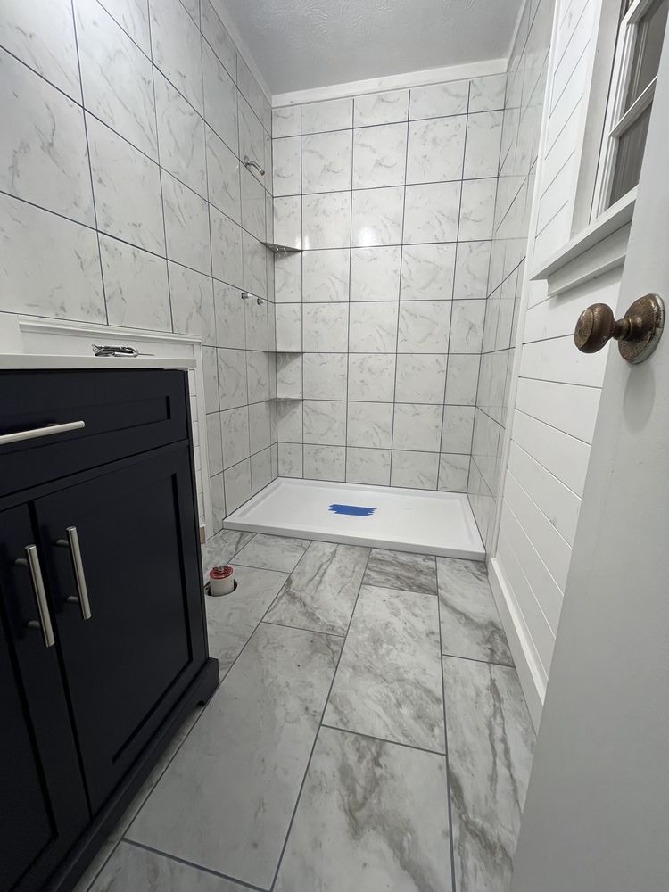 Bathroom with marble-look tiles. Dark blue vanity on left. Shower stall with two shelves and white base.