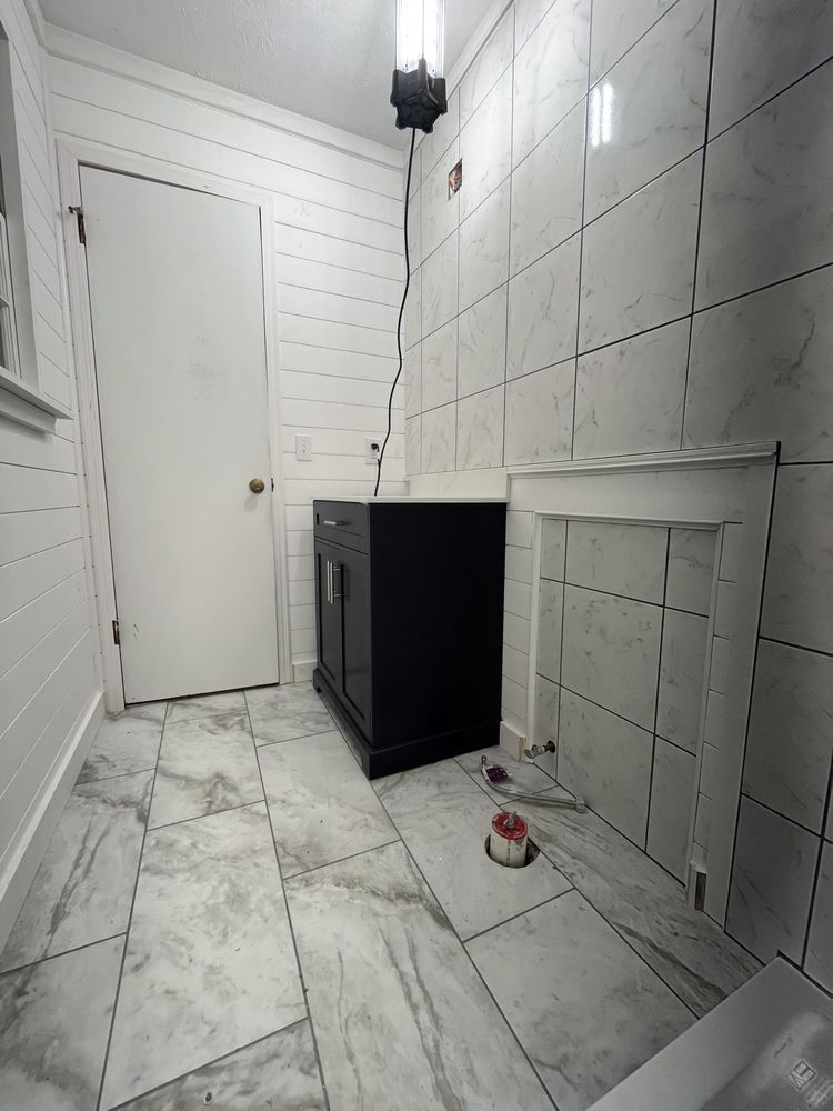Bathroom with marble-look floor and tiled wall. A dark vanity, white door, and unfinished wall section are visible.