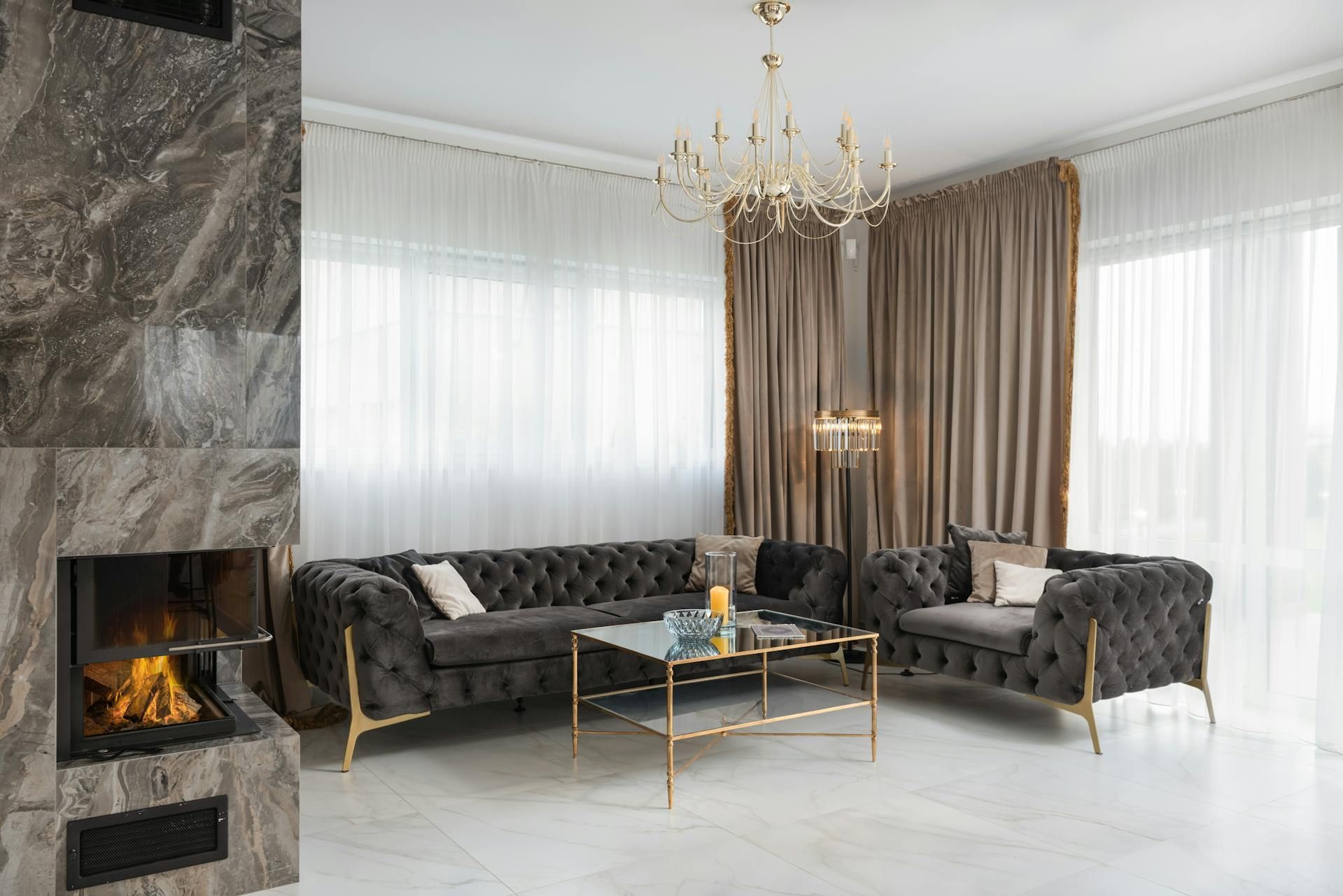 Living room with tufted gray velvet sofa and armchair, marble fireplace, and gold chandelier.