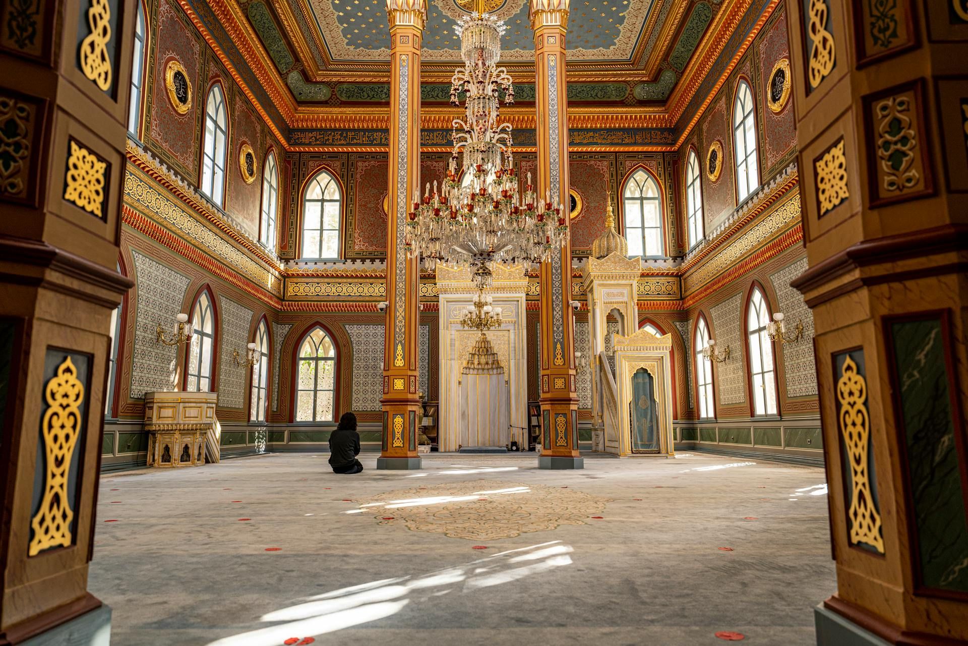 Ornate interior of a mosque with a person kneeling in prayer. Gold accents, glass chandelier, and window light.