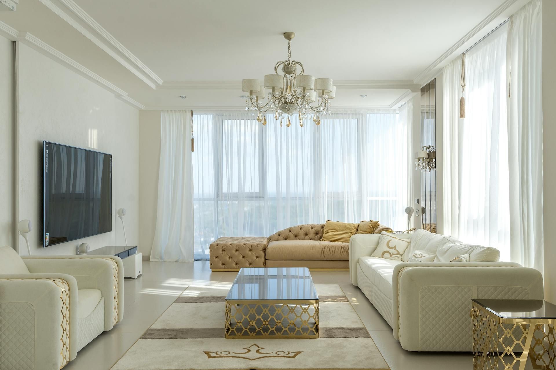 Elegant white living room with beige sofas, a glass table, and a chandelier, illuminated by sunlight.