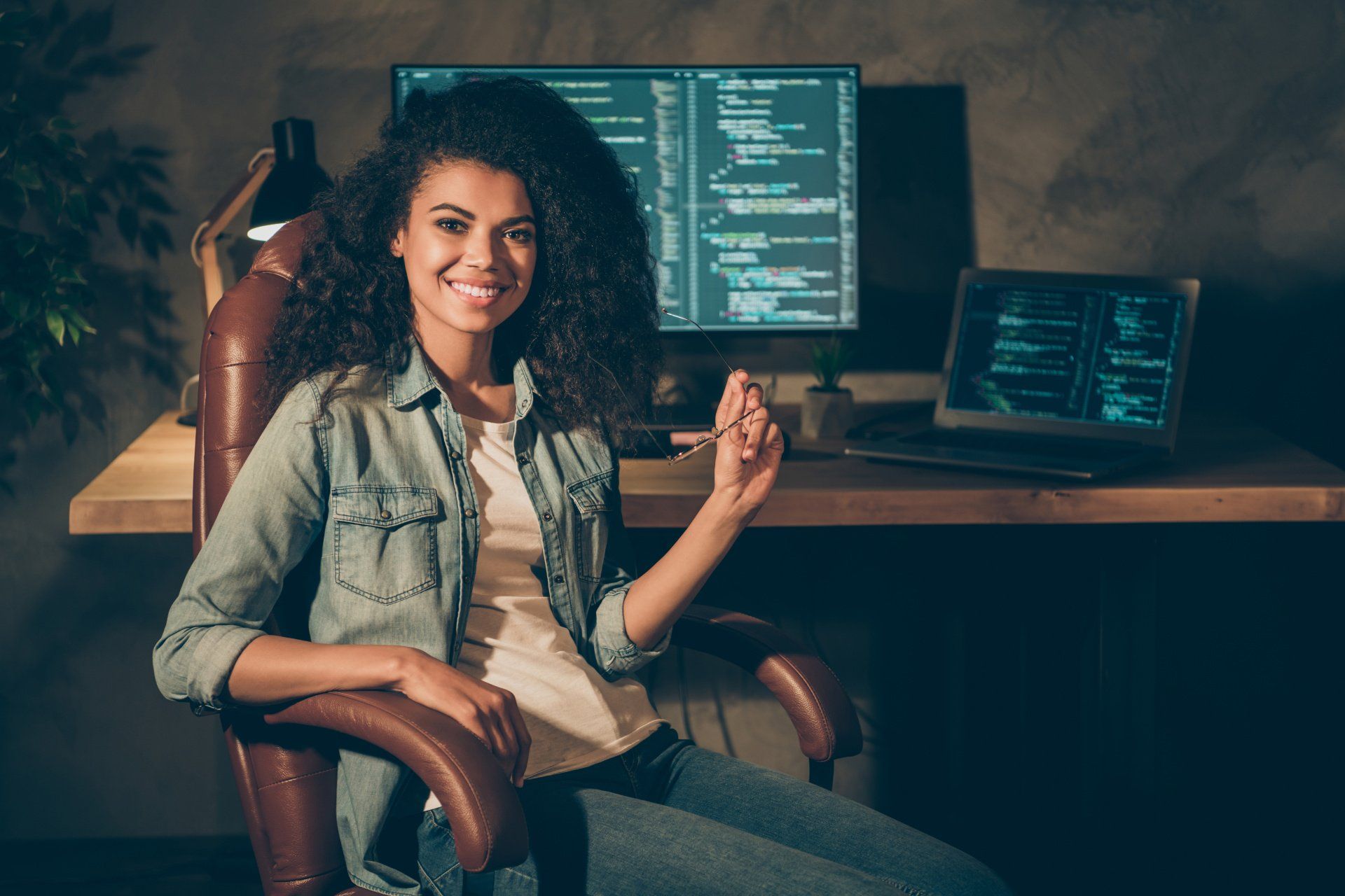 Woman with curly hair holding eye glasses with computer monitors behind her showing code