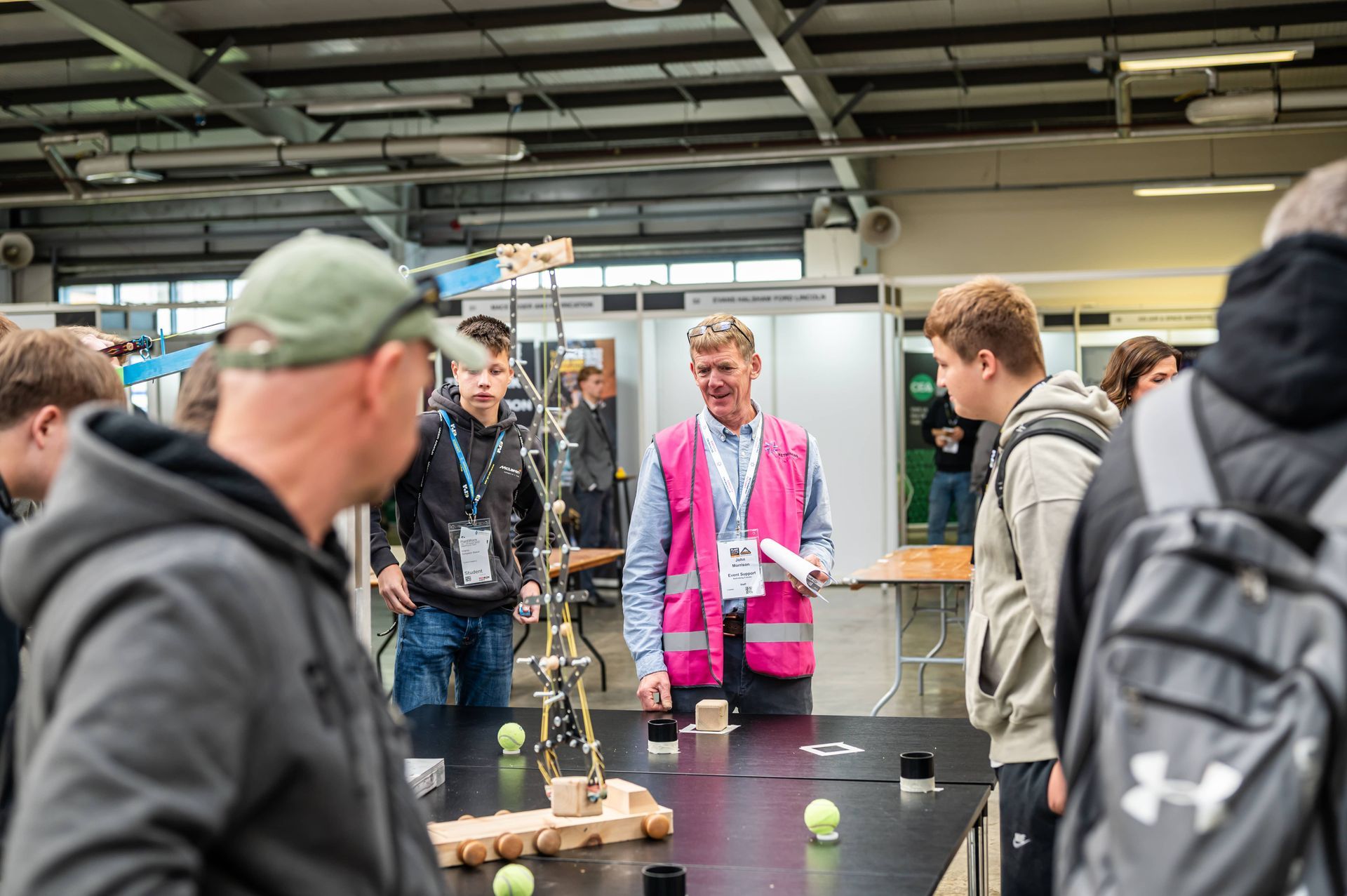 People observe a STEM project. A man in pink vest speaks while others look on. Indoor event.