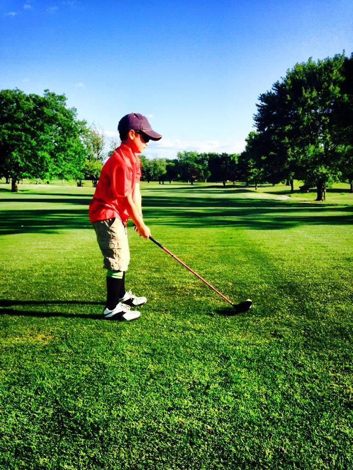 Boy in red shirt and cap swings golf club on a green course under a blue sky.
