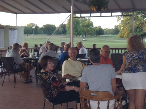 People seated at tables on a patio overlooking a golf course. Some are talking and looking at each other.