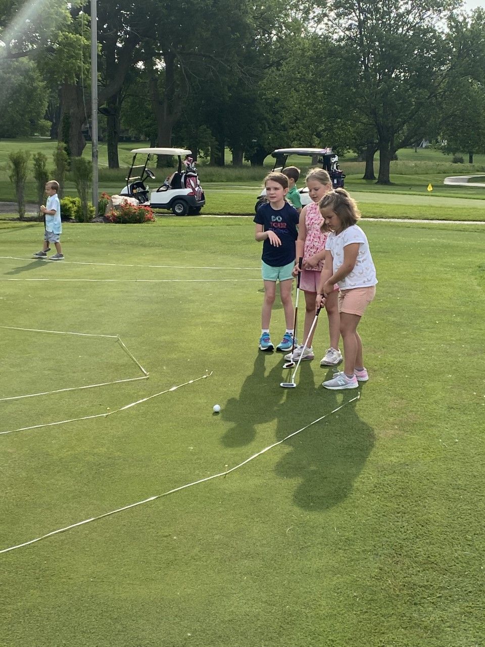 Four children putting on a green golf course, one child watches. Golf carts and trees in the background.