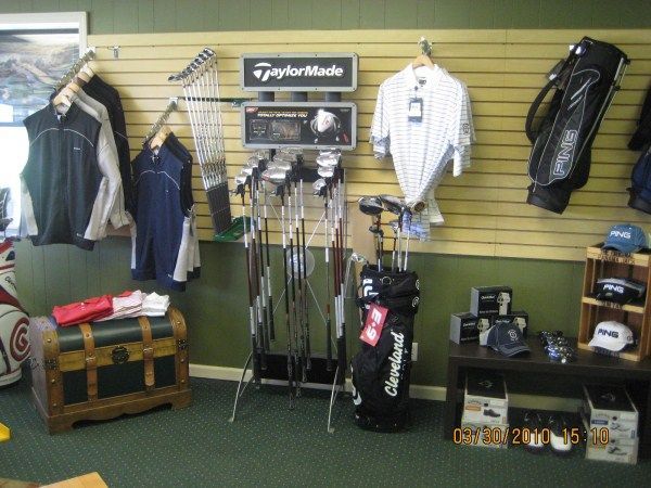 Golf shop interior with clubs, apparel, bags, and TaylorMade display. Green carpet, wood shelving, and a trunk.