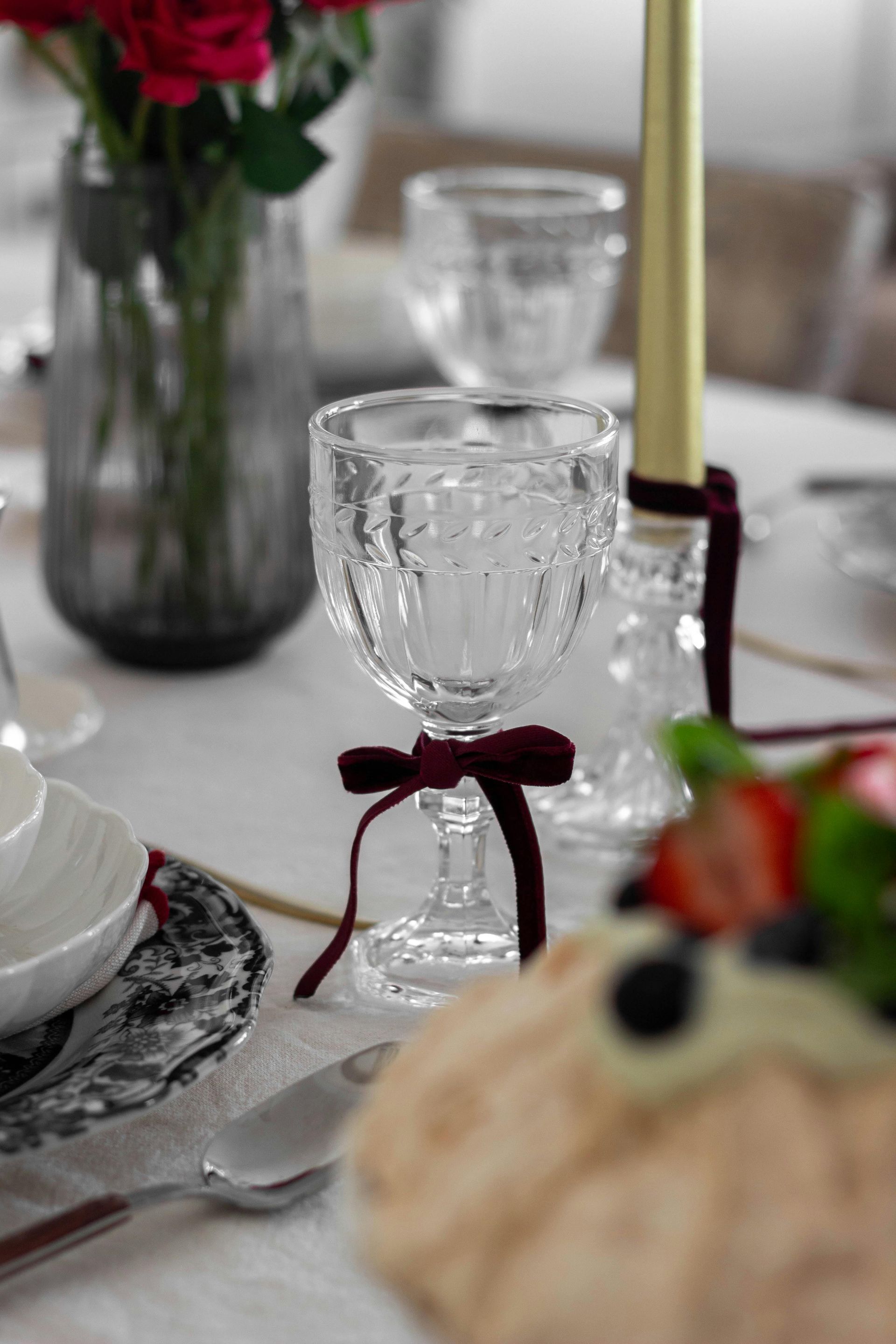 Crystal goblet with burgundy velvet ribbon, set on a white tablecloth; table setting with flowers and cake.