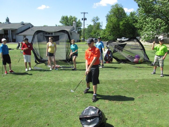 People practicing golf swings on a green, with nets in the background and a sunny sky.
