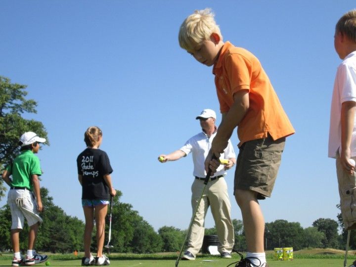 Children learning golf with an instructor on a green field under a clear blue sky.