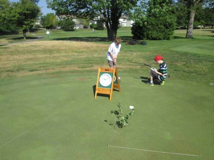 Two children playing golf on a green; one is about to putt, the other watches. A wooden scorecard sign is in view.