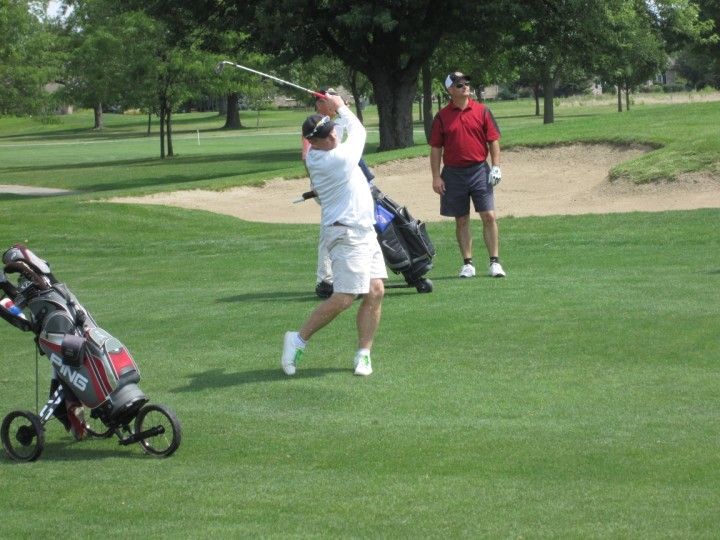 Golfer swings club on green, another watches. Golf bag nearby. Sunny day.