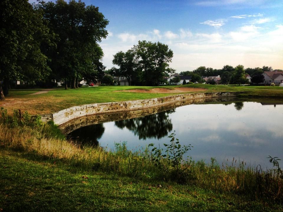 Pond with a stone retaining wall, green grass, trees, and a cloudy blue sky.
