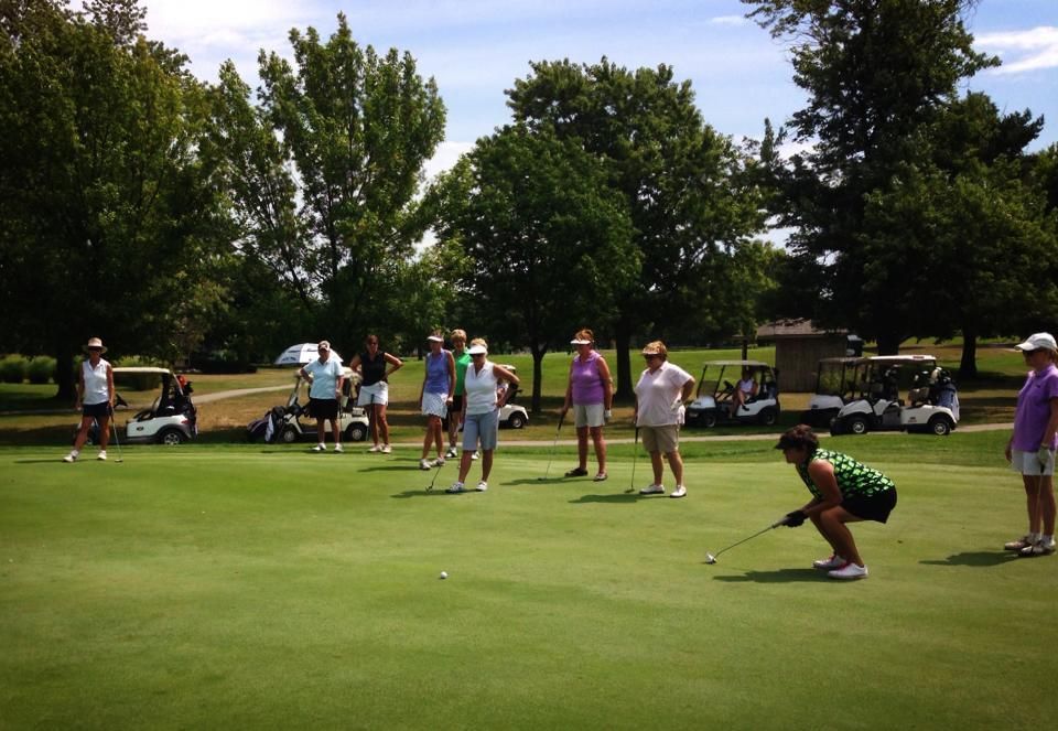Golfers putting on a green, sunny day. Several stand and watch. Golf carts and trees in the background.