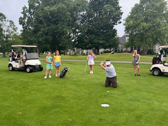 Golfers on a driving range under tents, lined up to hit balls. Overcast day.