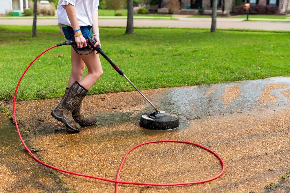 A Woman is Using a Pressure Washer to Clean a Driveway — Exquisite Exterior Cleaning In Terrigal, NSW