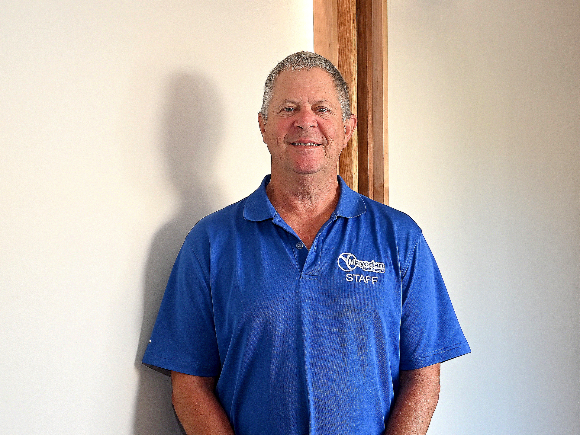 A man in a blue shirt is standing in front of a white wall.
