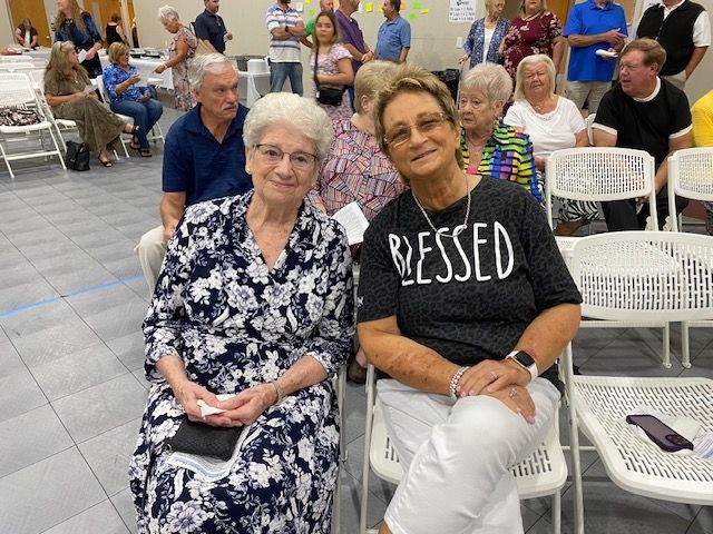 A group of people are sitting in chairs in a room and one woman is wearing a blessed shirt.