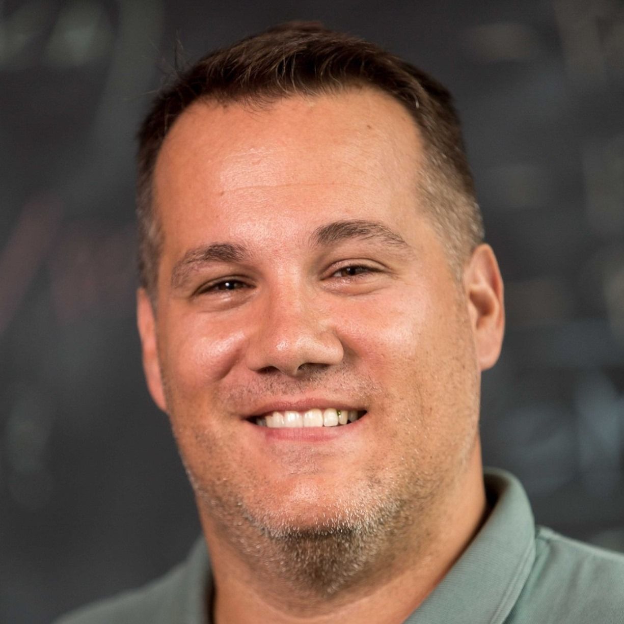 Moški s kratkimi rjavimi lasmi in svetlo brado se smeji, oblečen v zeleno polo majico.