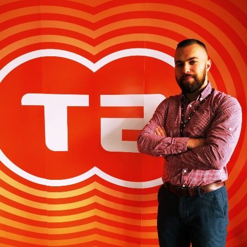 Moški z brado stoji pred rdečim logotipom s prekrižanimi rokami, oblečen v vzorčasto majico in temne hlače.