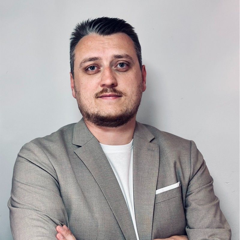 Moški s kratkimi temnimi lasmi v rjavorjavem suknjiču, beli srajci, s prekrižanimi rokami, gleda v kamero. Belo ozadje.