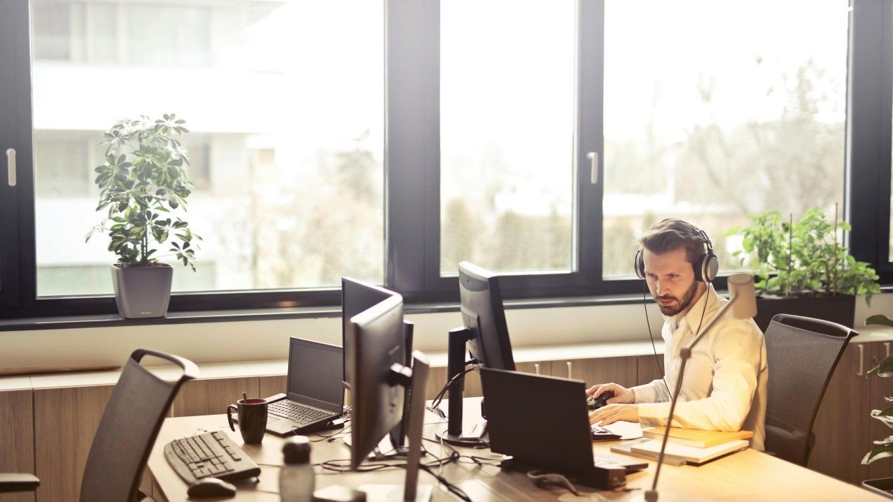 Man in office with headphones, working on computer. Brightly lit with window view and plants.