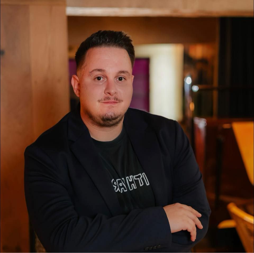 Moški v črnem suknjiču s prekrižanimi rokami, nasmejan, v zaprtih prostorih z lesenimi oblogami in restavracijskim vzdušjem.