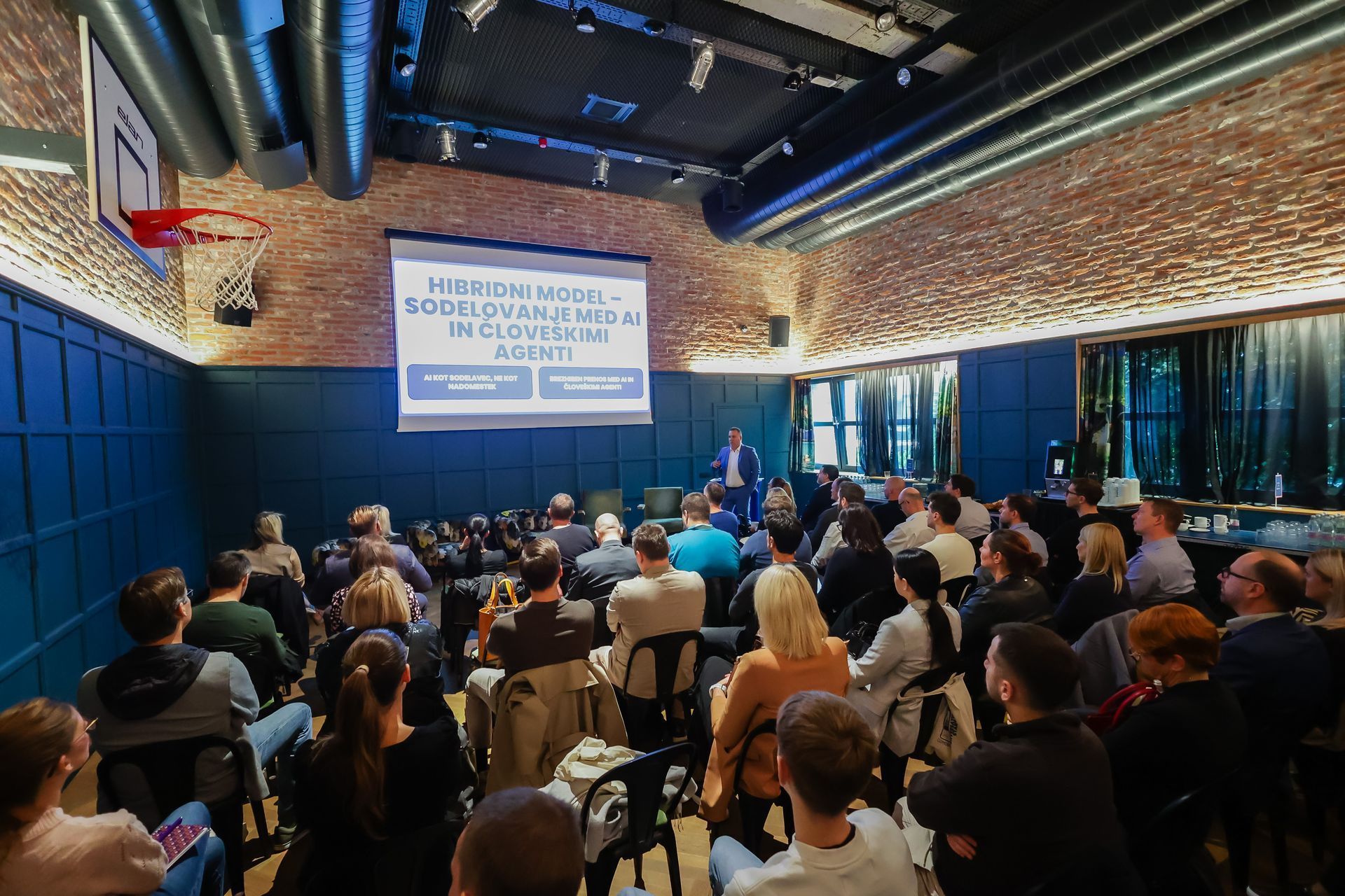 Občinstvo sedi in posluša predstavitev na odru; notranjost v modri in opečnati barvi.