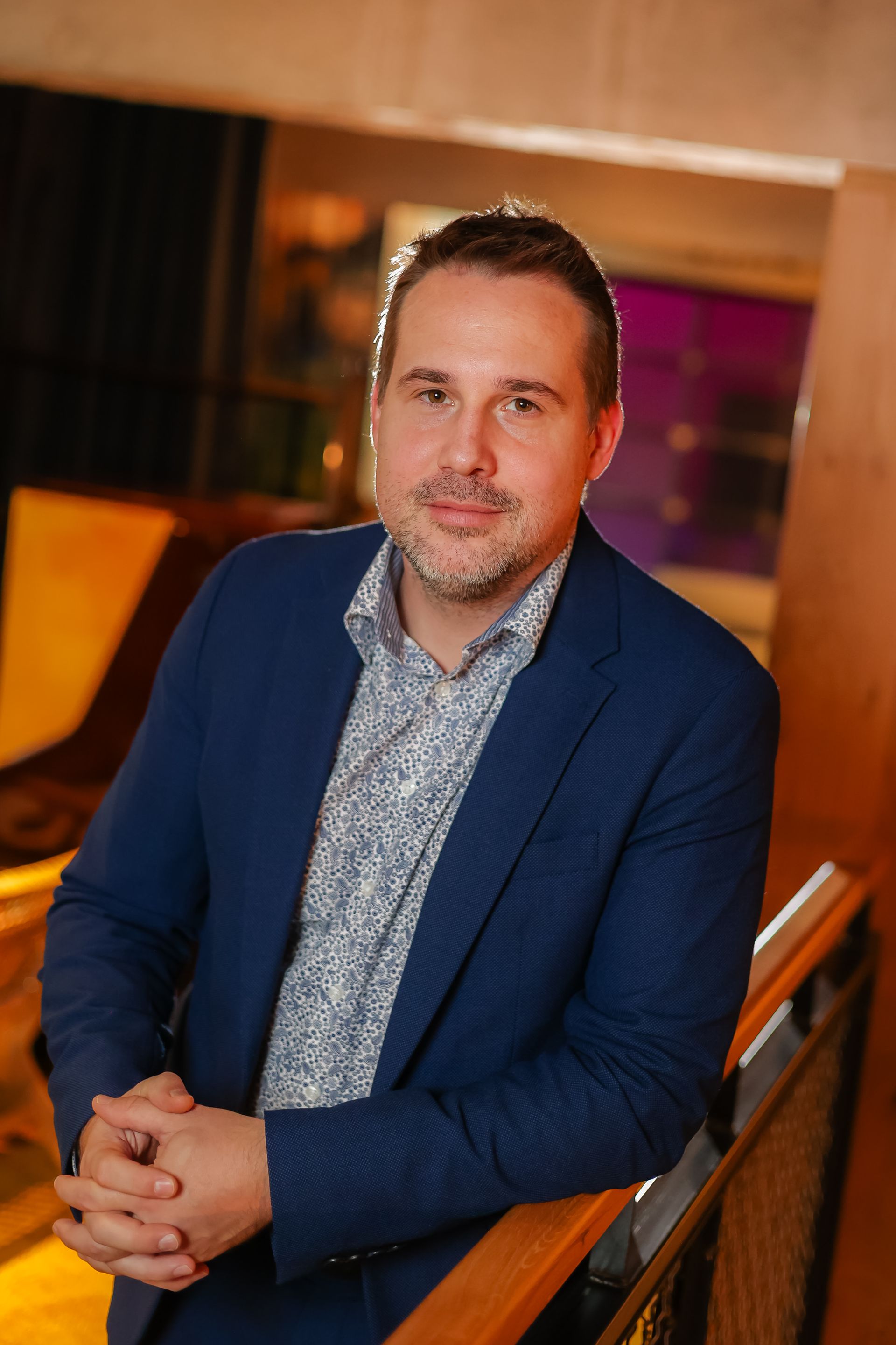 Moški v modrem suknjiču in vzorčasti srajci se naslanja na ograjo in se rahlo nasmehne. Zamegljeno ozadje notranjosti.