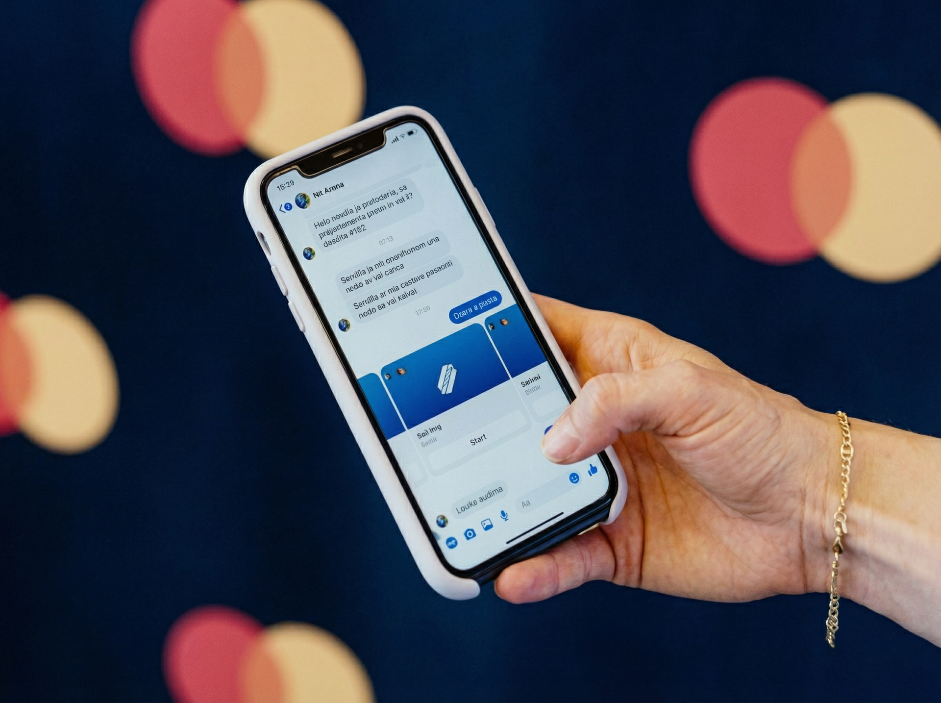 Roka drži bel telefon, ki prikazuje aplikacijo za sporočanje na modrem ozadju z rdečimi in bež krogi.