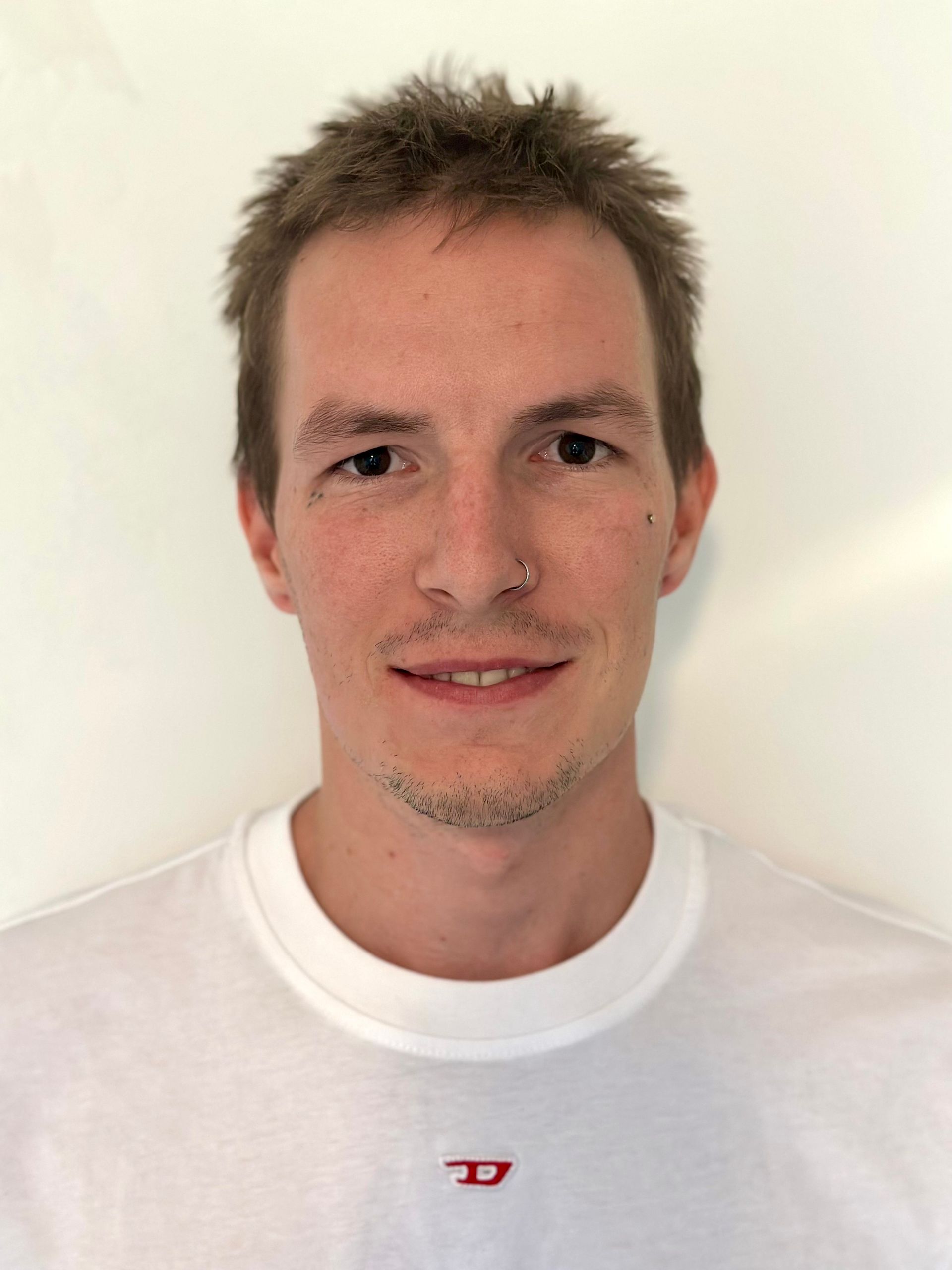 Moški s kratkimi lasmi in obročkom v nosu se rahlo nasmehne. Oblečen je v belo srajco ob beli steni.