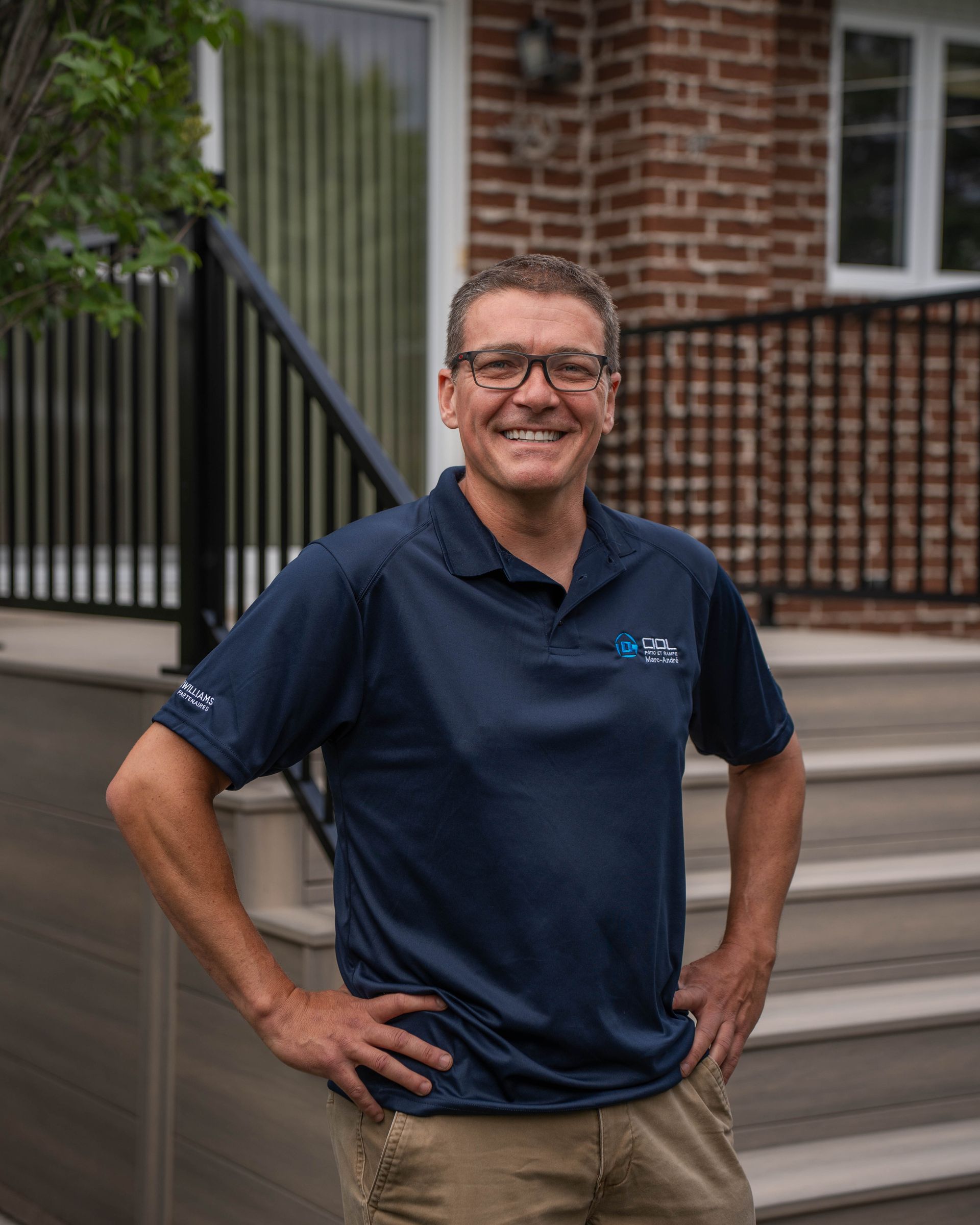 Un homme portant des lunettes et une chemise bleue se tient sur un escalier devant une maison en briques.
